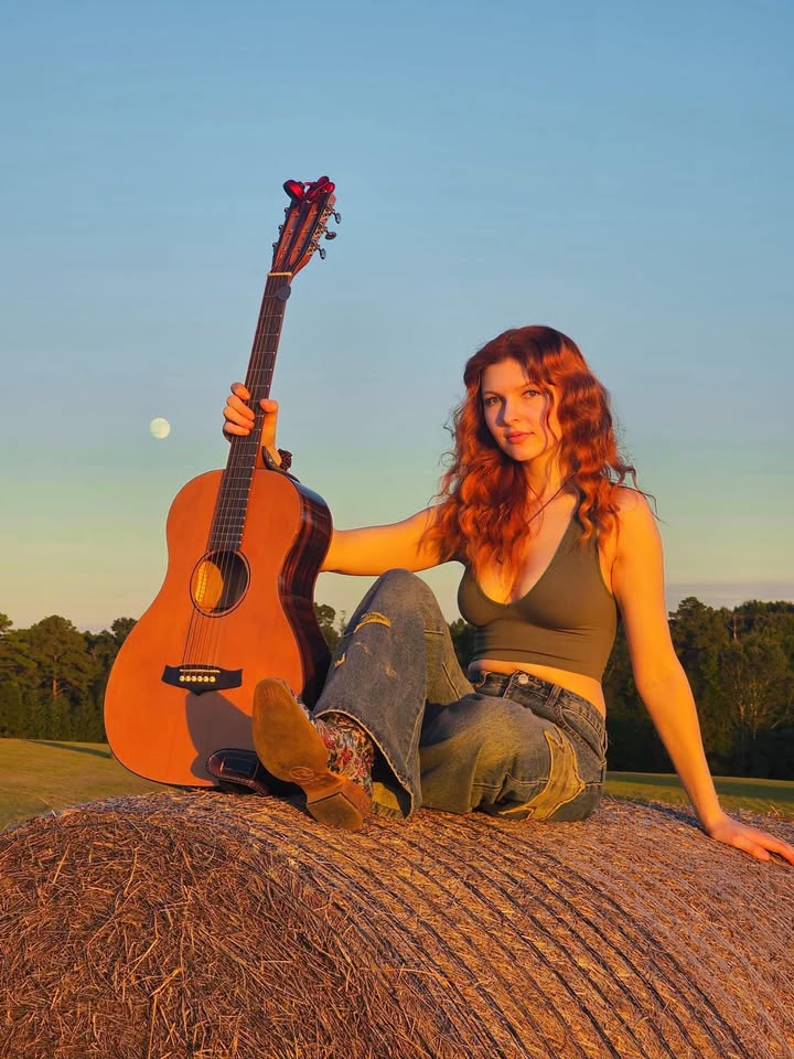 Caitlin Mae sitting on a hay bale at sunset with her acoustic guitar